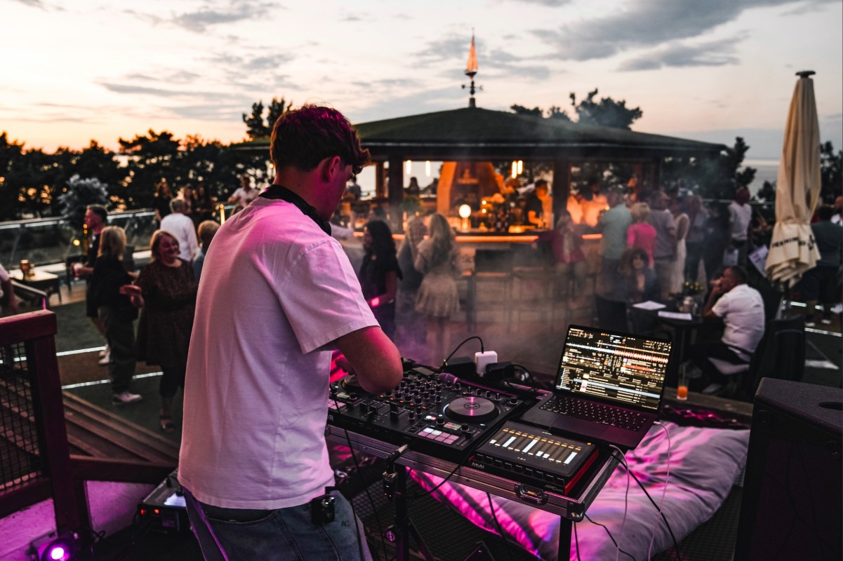 DJ legt bei Sonnenuntergang auf der Sky Bar Binz auf, im Hintergrund tanzende Gäste unter der überdachten Bar-Pergola mit Blick auf die Ostsee
