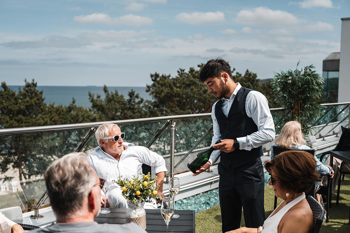 Servicekraft öffnet eine Flasche Sekt am Tisch der Sky Bar in Binz mit Blick auf die Ostsee.
