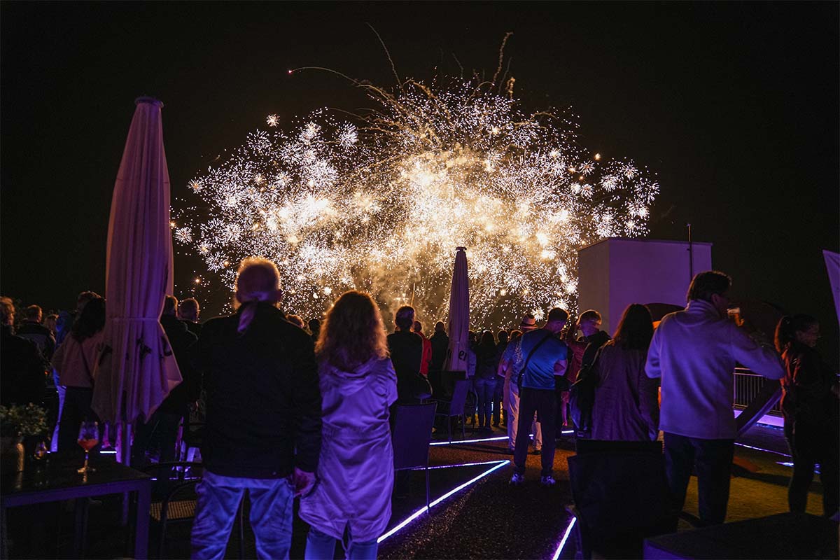 Gäste auf der nächtlichen Dachterrasse der Sky Bar Binz beobachten ein goldenes Feuerwerk