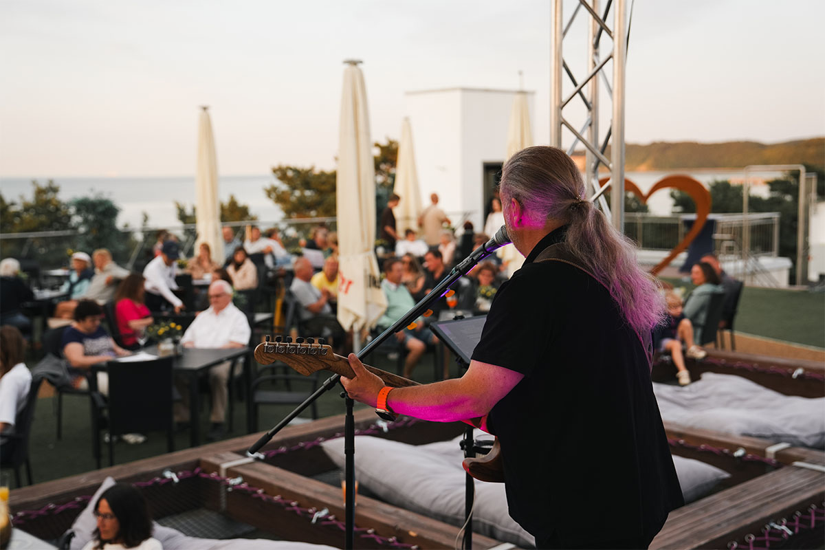 Gitarrist spielt auf der Dachterrasse der Sky Bar Binz vor Gästen an Tischen und Netzliegen, im Hintergrund die Ostsee