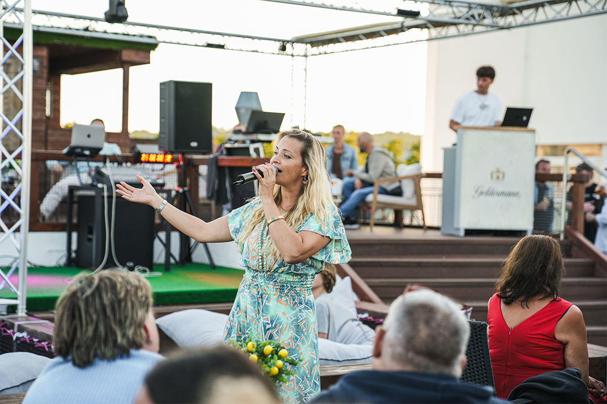 Sängerin mit Mikrofon singt zwischen den Gästen auf der Dachterrasse der Sky Bar Binz, im Hintergrund das DJ-Pult