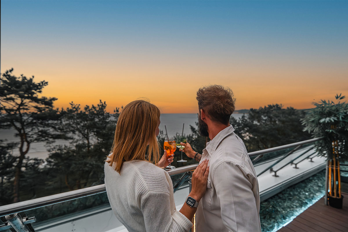 Paar stößt auf der Terrasse der Arkona Sky Bar mit Cocktails an, im Hintergrund Sonnenuntergang und Meerblick